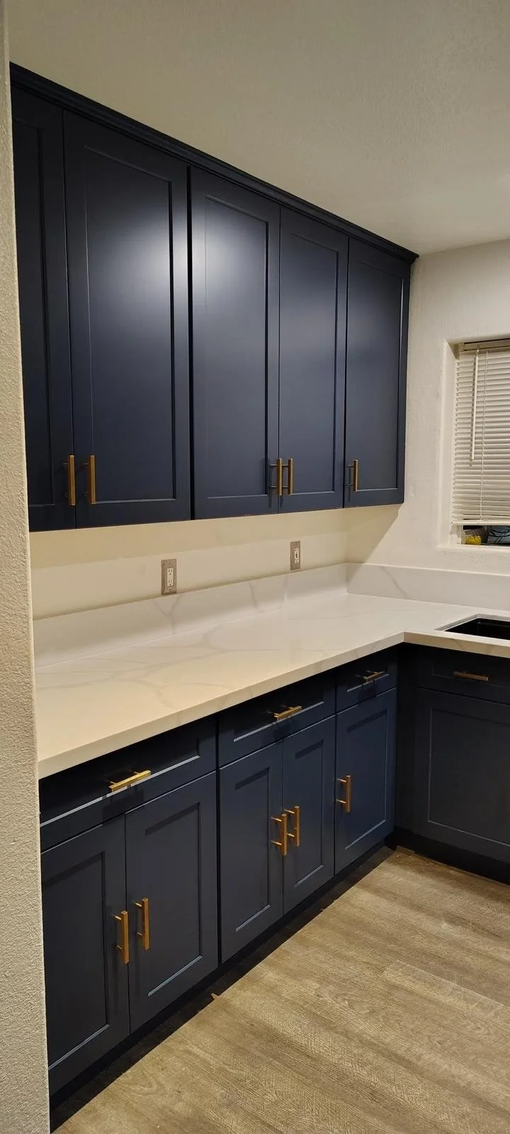 Kitchen with navy blue cabinets, white marble countertop, beige walls, and a small window with blinds.