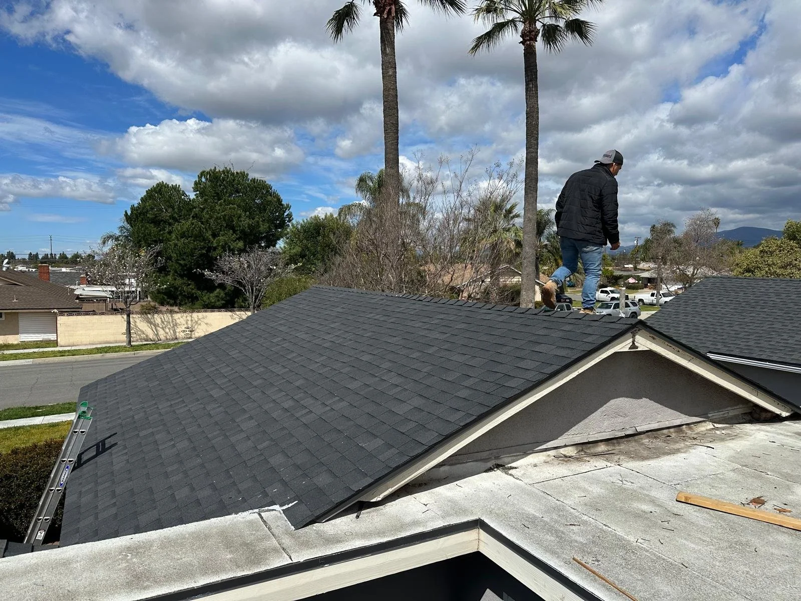 A man standing on a roof working on shingles, with a ladder leaning against the roof, surrounded by trees and houses under a partly cloudy sky.