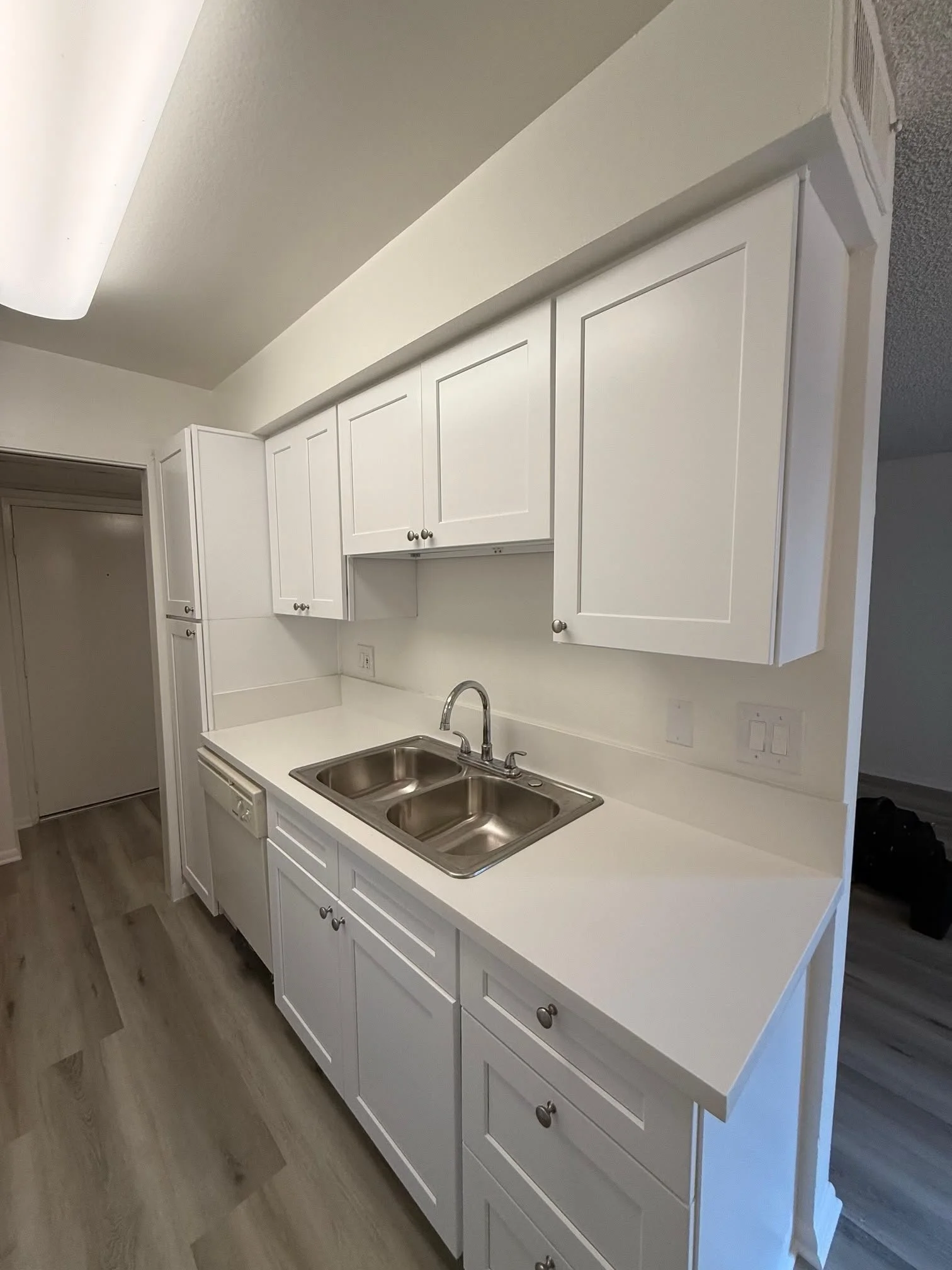 Kitchen with white cabinets, stainless steel double sink, beige countertops, wood flooring, and a ceiling light fixture.