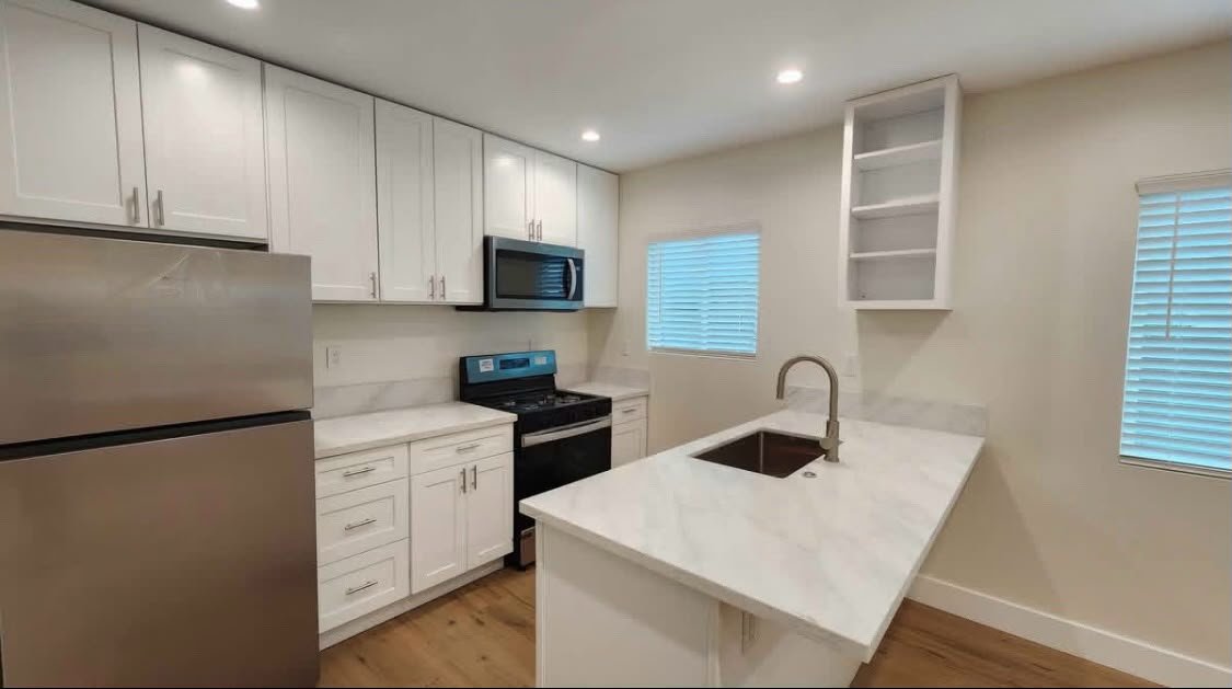 Modern kitchen with white cabinets, stainless steel refrigerator, microwave, and stove, marble countertops, small open shelving, two windows with blinds, and a kitchen island with a sink.