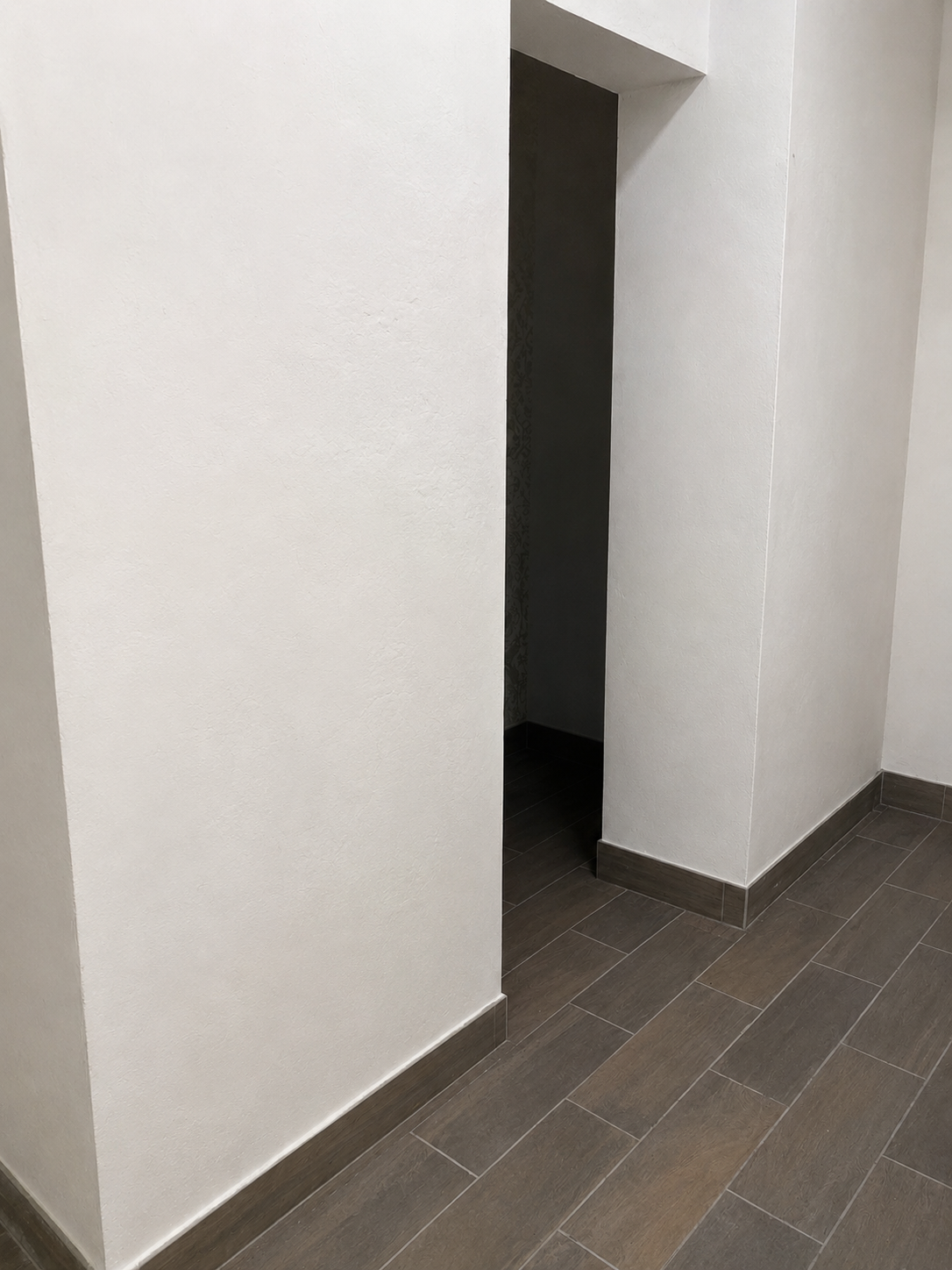 A hallway corner with white textured walls, dark brown floor tiles, and a small entrance to a dark room or closet.