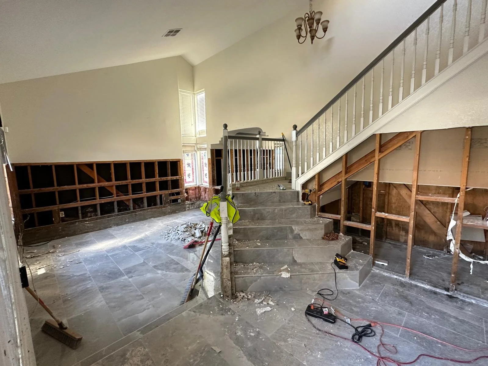 Living room under renovation with torn linoleum, exposed wall framing, tools, and construction debris, featuring a staircase with white railing and a chandelier.