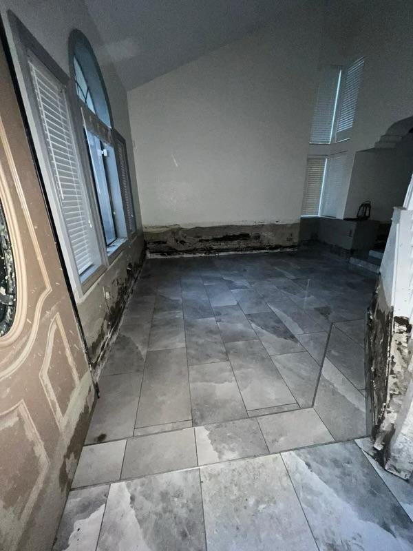 Room with partially completed tiled floor, peeling paint and damage around base of walls, and windows with blinds.