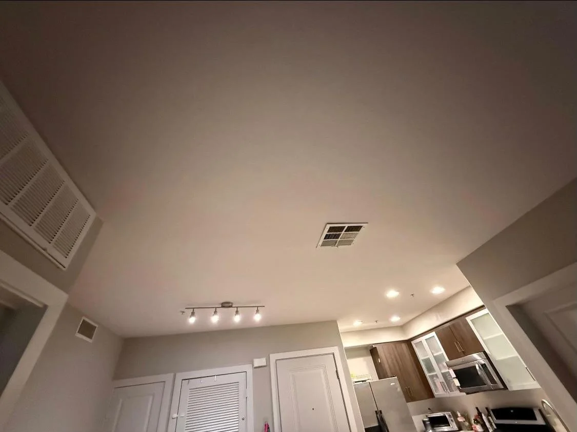 Living room ceiling with ceiling vent, track lighting, and recessed lights, with kitchen in background.