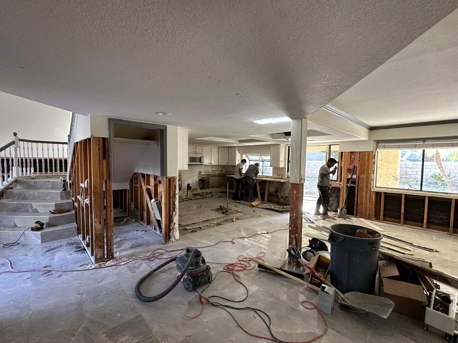 Interior of a house under renovation with workers demolishing walls, exposed wooden framing, construction debris, tools, and a large trash bin.