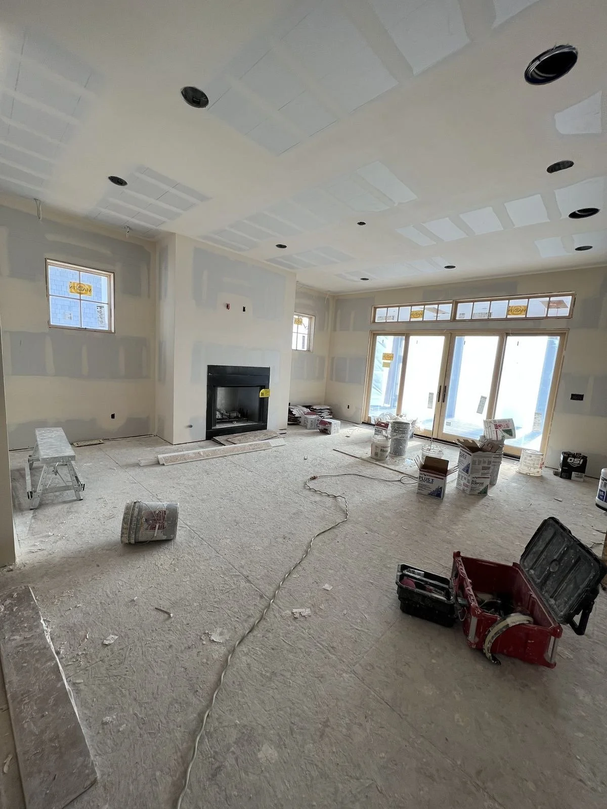 Living room under construction with drywall installed and ceiling lights being prepared, windows and sliding glass door open to outside, construction materials and tools scattered on unfinished floor.