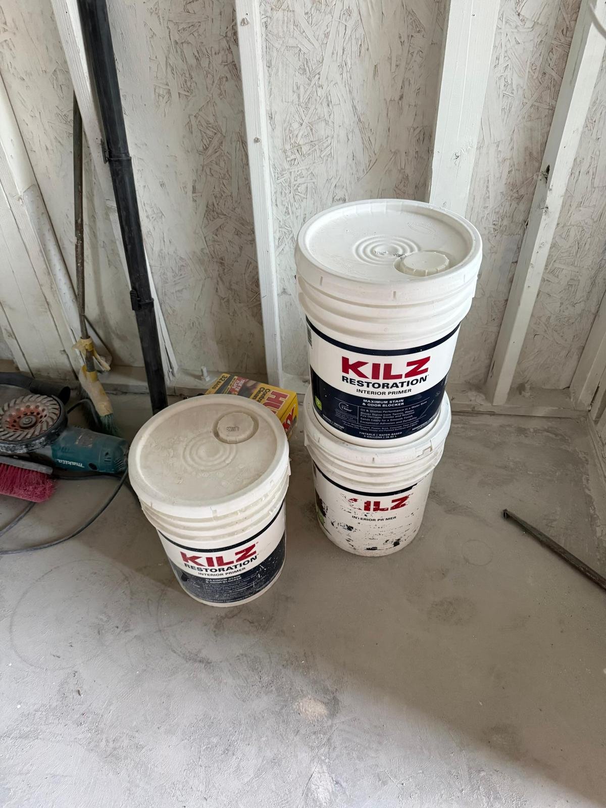Three buckets of Kilz Restoration interior primer stacked on a concrete floor in a room under construction with drywall framing, a window, and construction tools nearby.