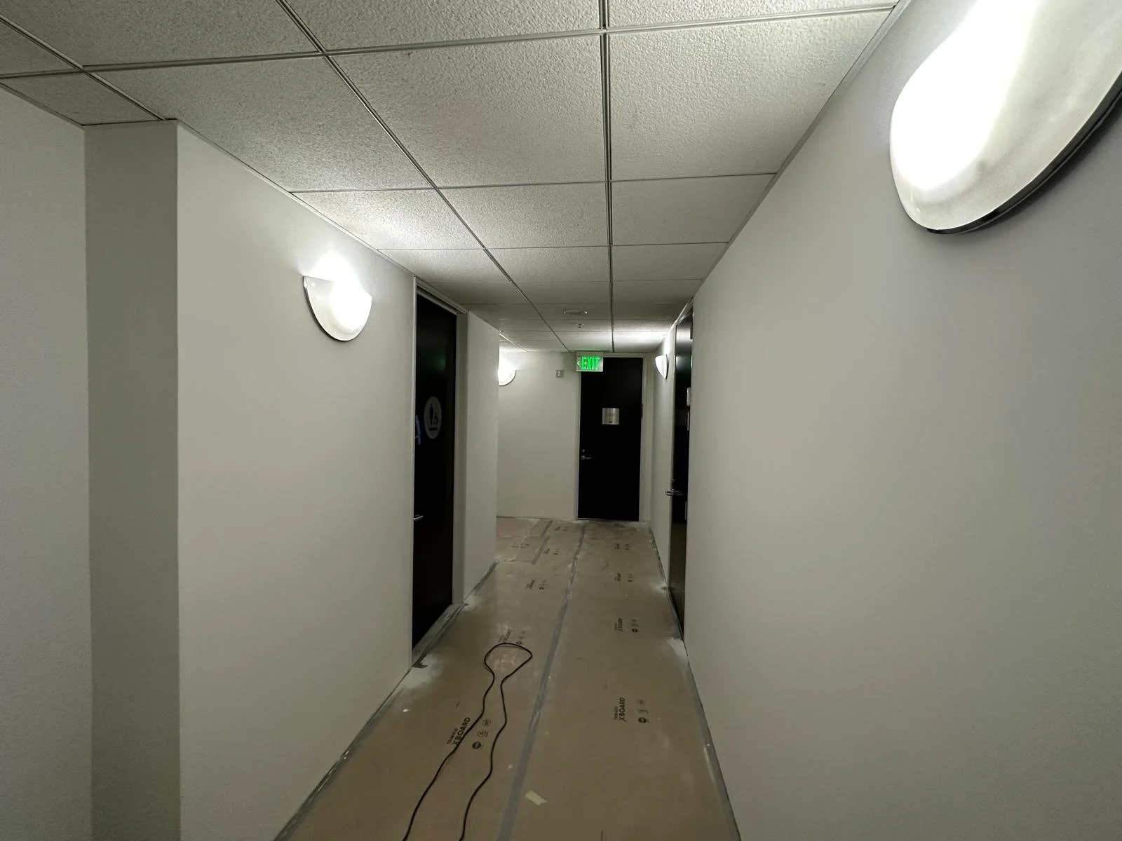 An indoor hallway with white walls, a beige floor, and ceiling lights, leading to a black door with a green exit sign above it.