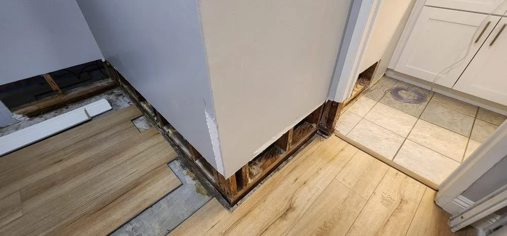 A kitchen corner with a partially removed baseboard, exposing the wooden wall studs and subflooring, with new flooring being installed.