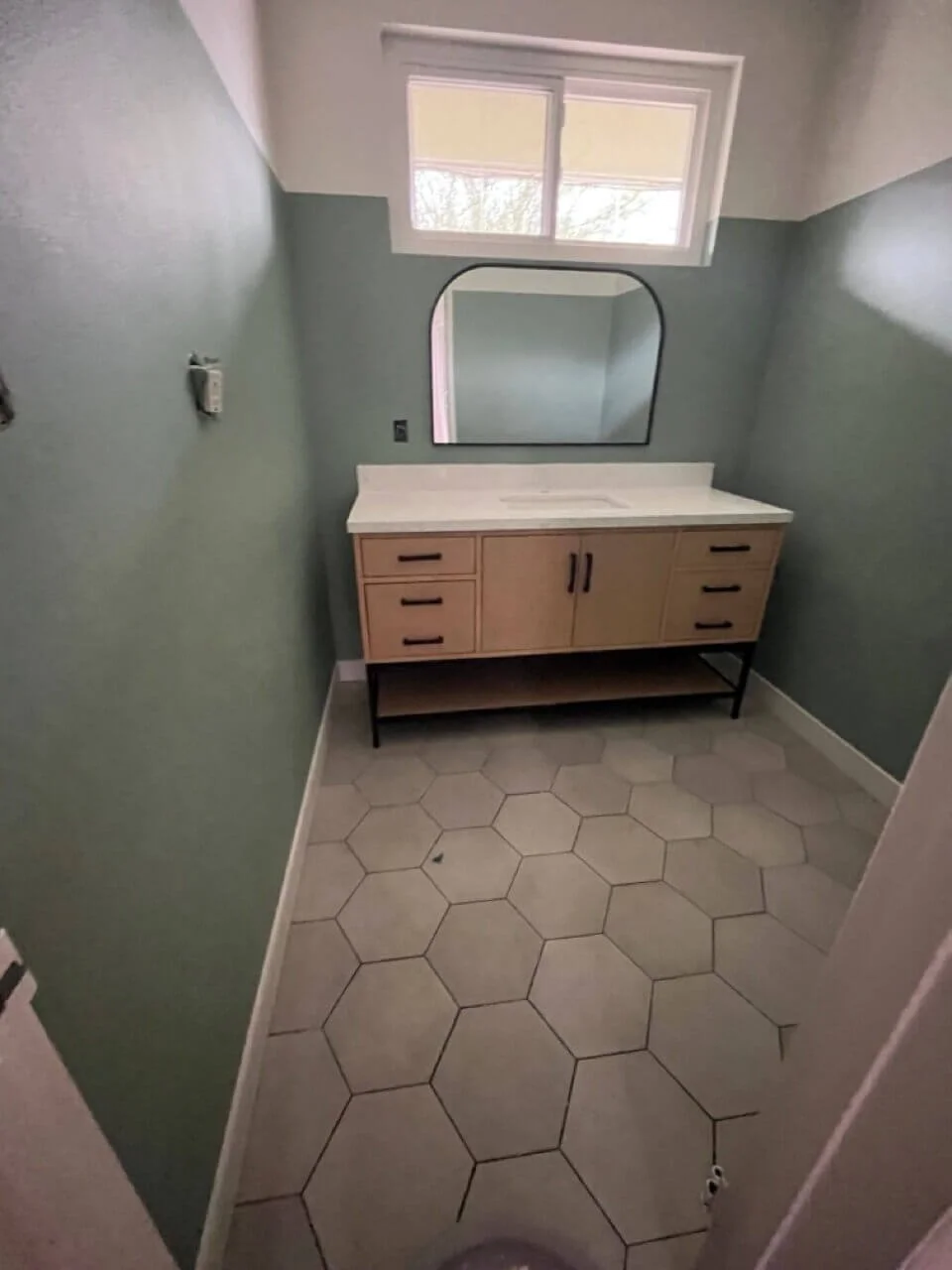 Empty bathroom with hexagonal tile floor, green walls, a wooden vanity with a mirror, and a small window