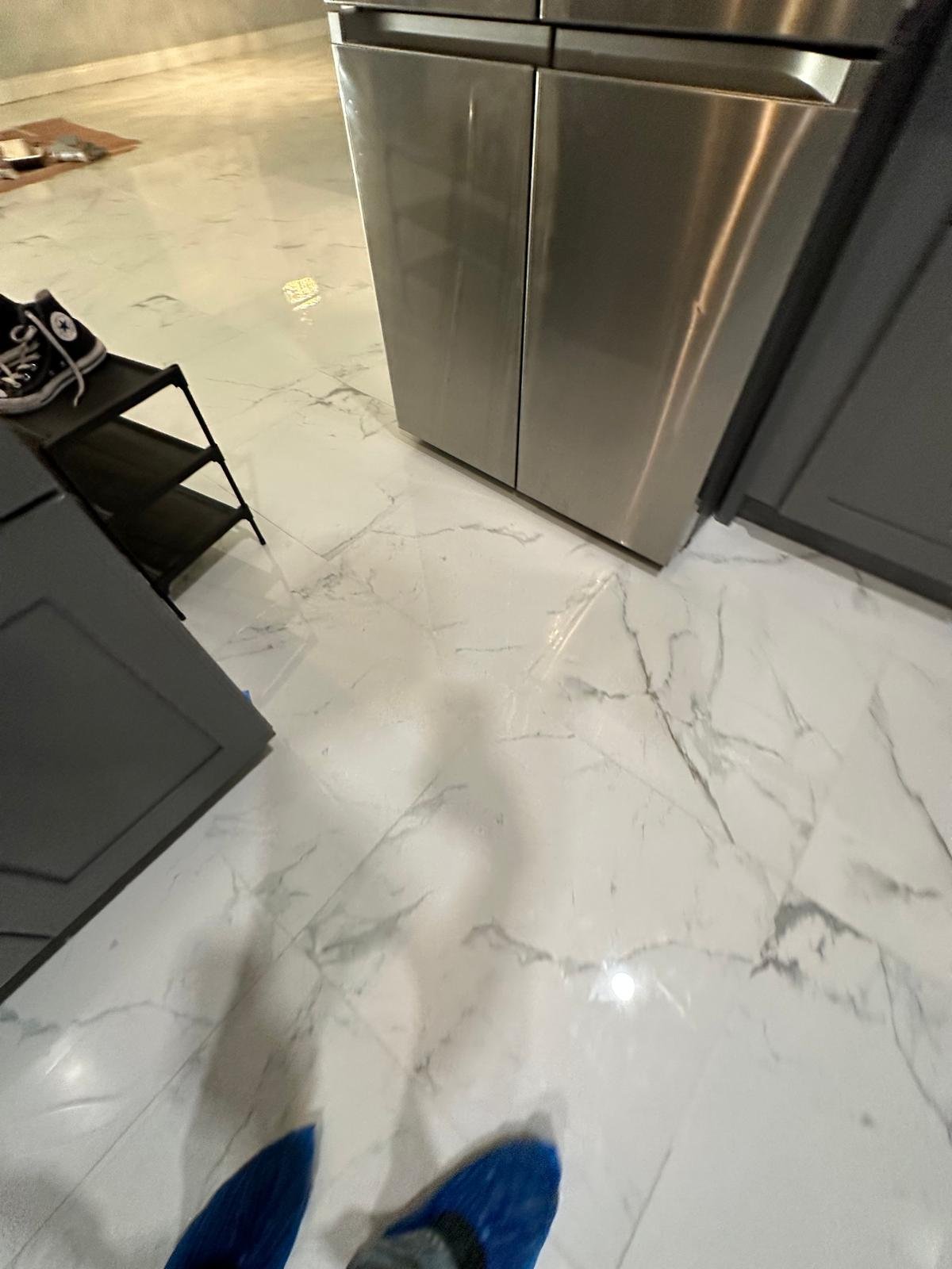 This image shows a part of a kitchen with glossy white marble floors with gray veins. There are gray cabinets, a black shelving unit with sneakers on it, and a stainless steel appliance or cabinet.
