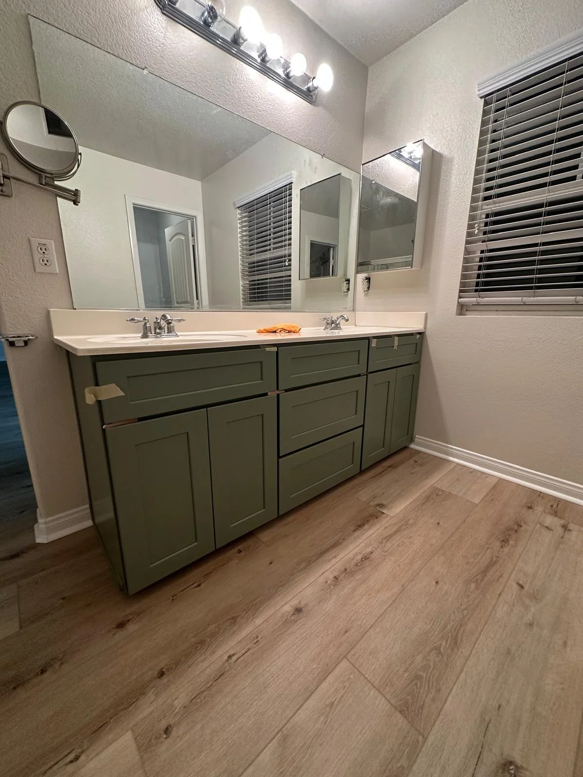 Bathroom vanity with green cabinets, a large mirror, and a four-light fixture above, with a window covered by blinds on the right wall.