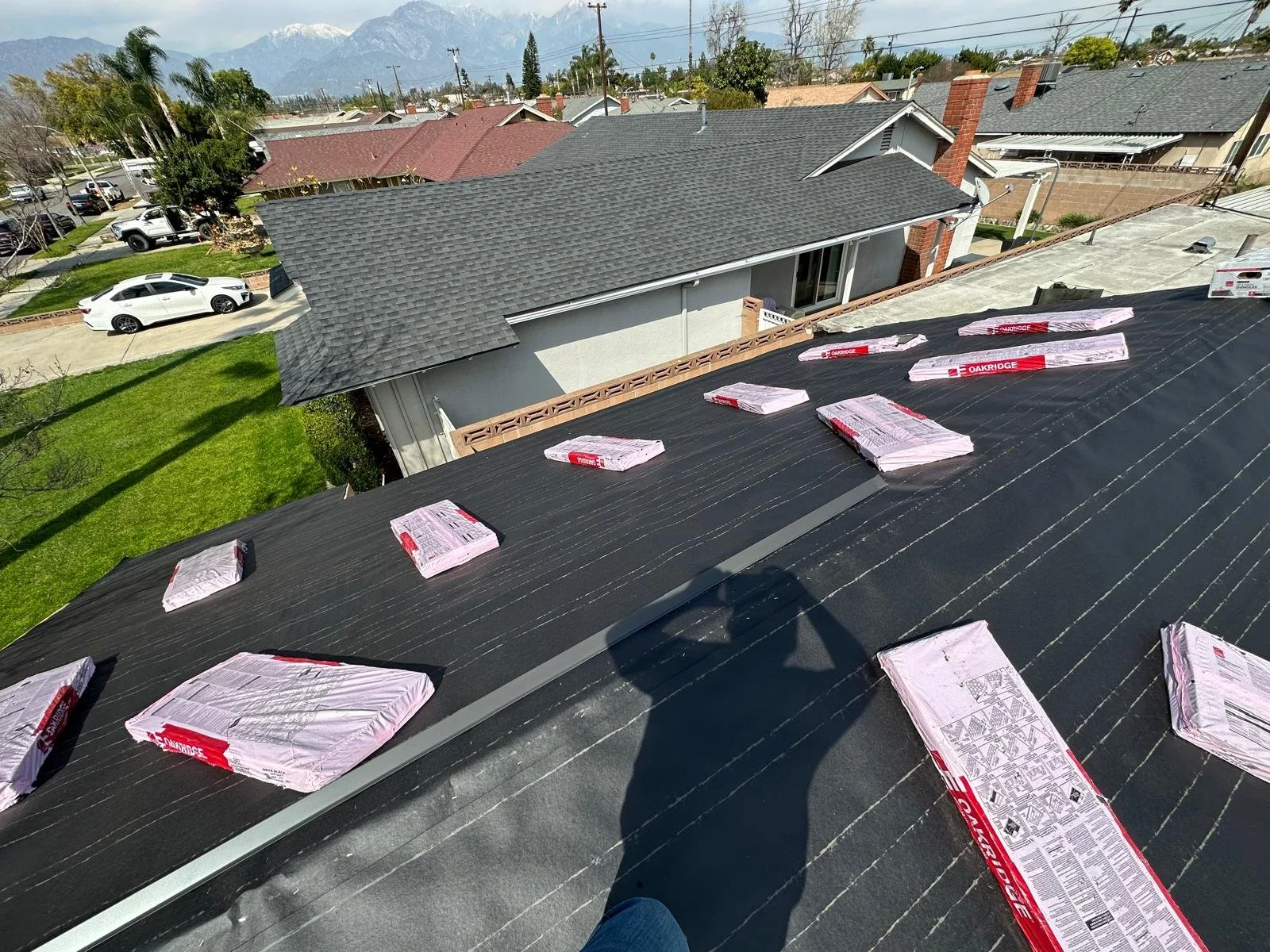 View from a rooftop under construction with shingles and roofing materials, residential houses, trees, parked cars, and distant mountains.