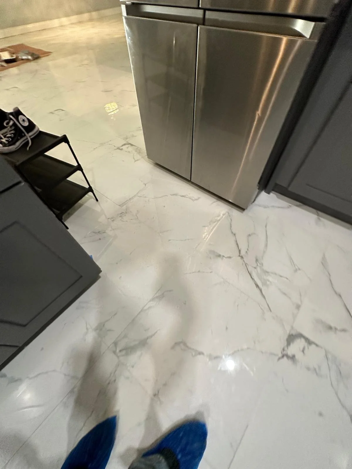View of a kitchen floor with white marble tiles, a stainless steel refrigerator, a black cabinet on one side, and a black shoe rack with shoes on top.