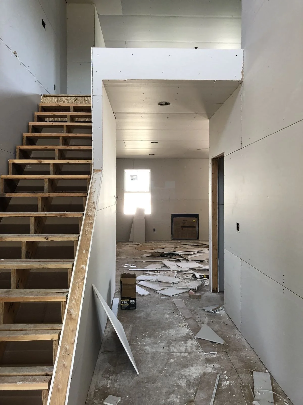 Interior of a house under construction with unfinished walls, wooden staircase, scattered construction debris, and a small window at the back.