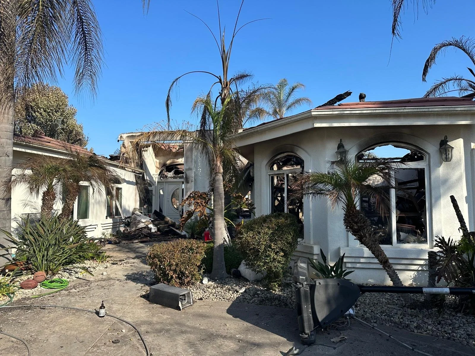 House with damaged roof and broken windows, debris on the ground, palm trees, and damaged garden after a fire or disaster.