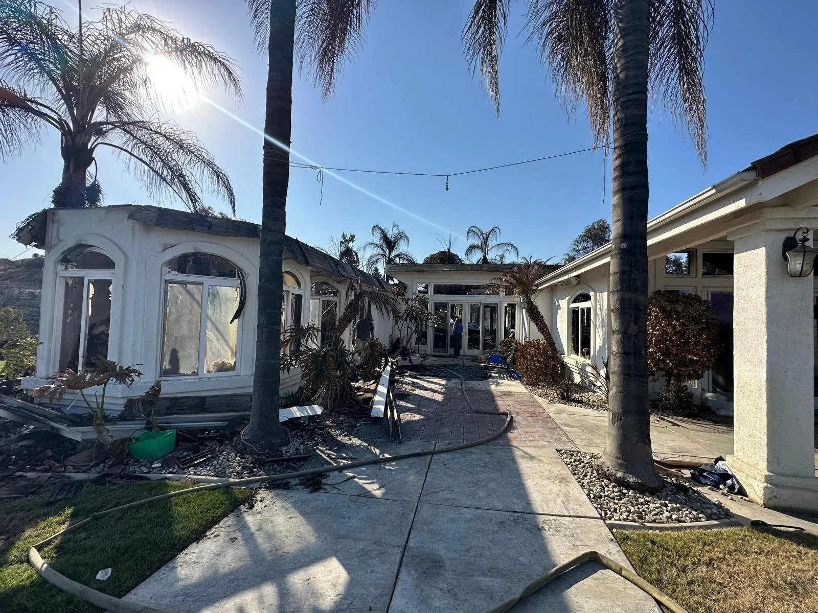 Damaged house with broken windows, palm trees, and debris scattered on a walkway, possibly after a storm or fire.