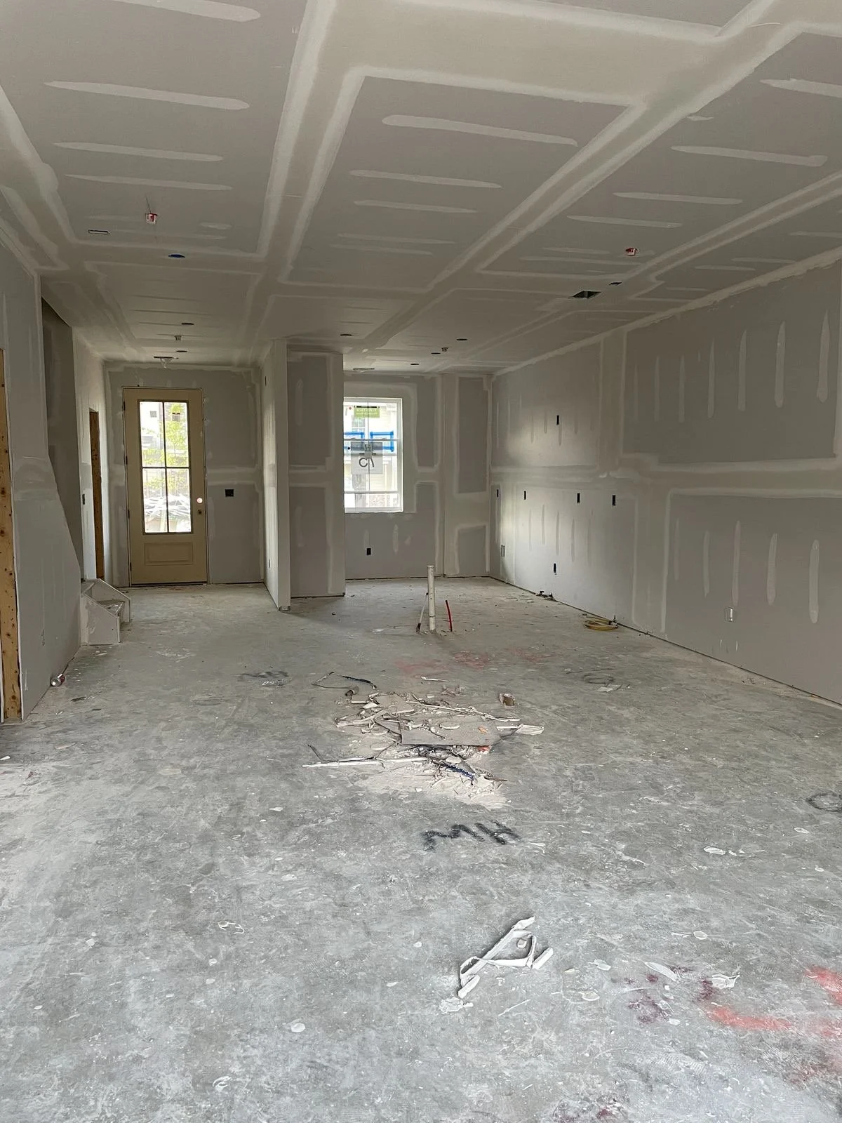 Interior of a house under construction with drywall partially installed on walls and ceiling, debris on the concrete floor, and a door and window at the back.