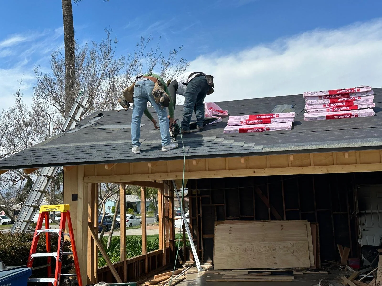 Two workers installing new asphalt shingles on the roof of a house under construction, with stacks of shingles labeled 'OAKRIDGE' nearby, on a clear day.