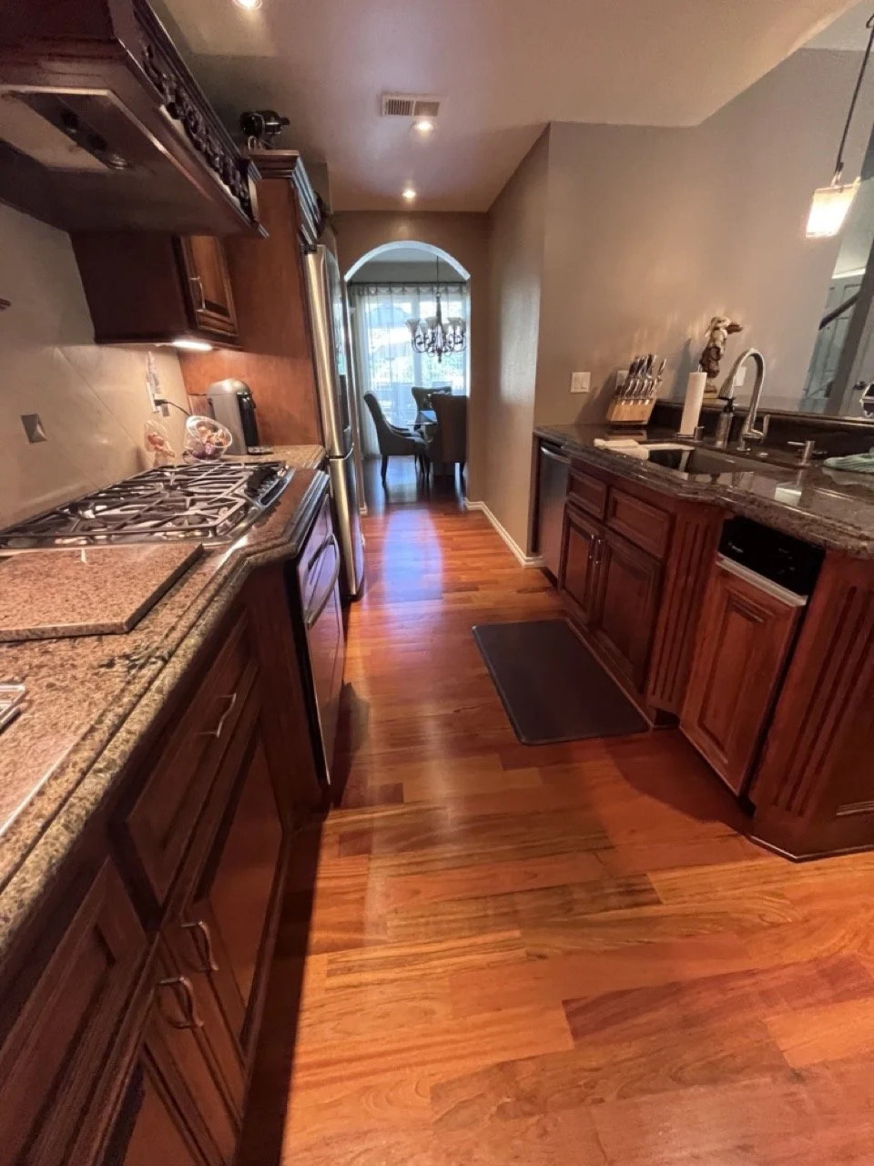 A narrow kitchen with wooden cabinets and drawers, a granite countertop, a stove, and stainless steel appliances. There is a sink with a faucet on the right side, and a small rug on the wooden floor. In the background, there is a dining area with a r