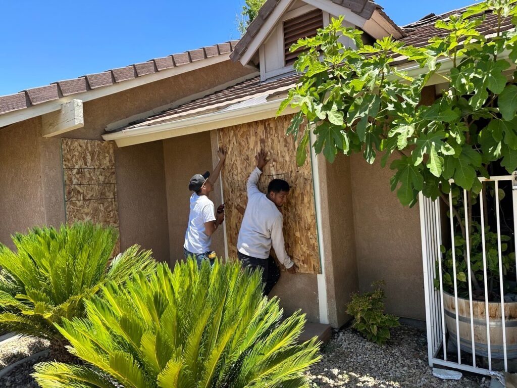 Two men are installing plywood on the exterior wall of a house next to a porch. One man is holding a screwdriver, and the other is positioning the plywood. There are green plants in the foreground and a tree partially covering the house. The house has a brown shingle roof.