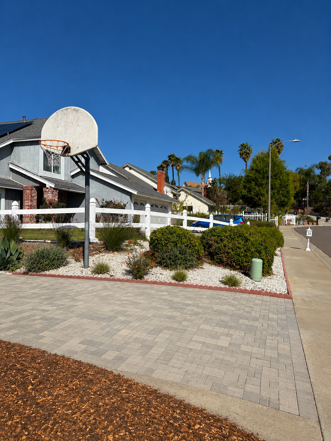 Residential neighborhood with a basketball hoop, house, palm trees, parked cars, sidewalk, street, and well-maintained garden with bushes and rocks.