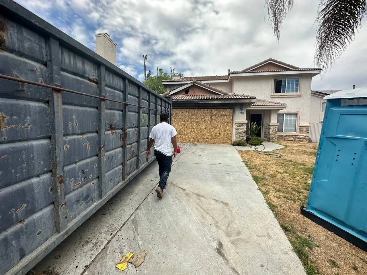 A person in a white shirt and dark jeans walking towards a house while pushing a large metal dumpster on the driveway.