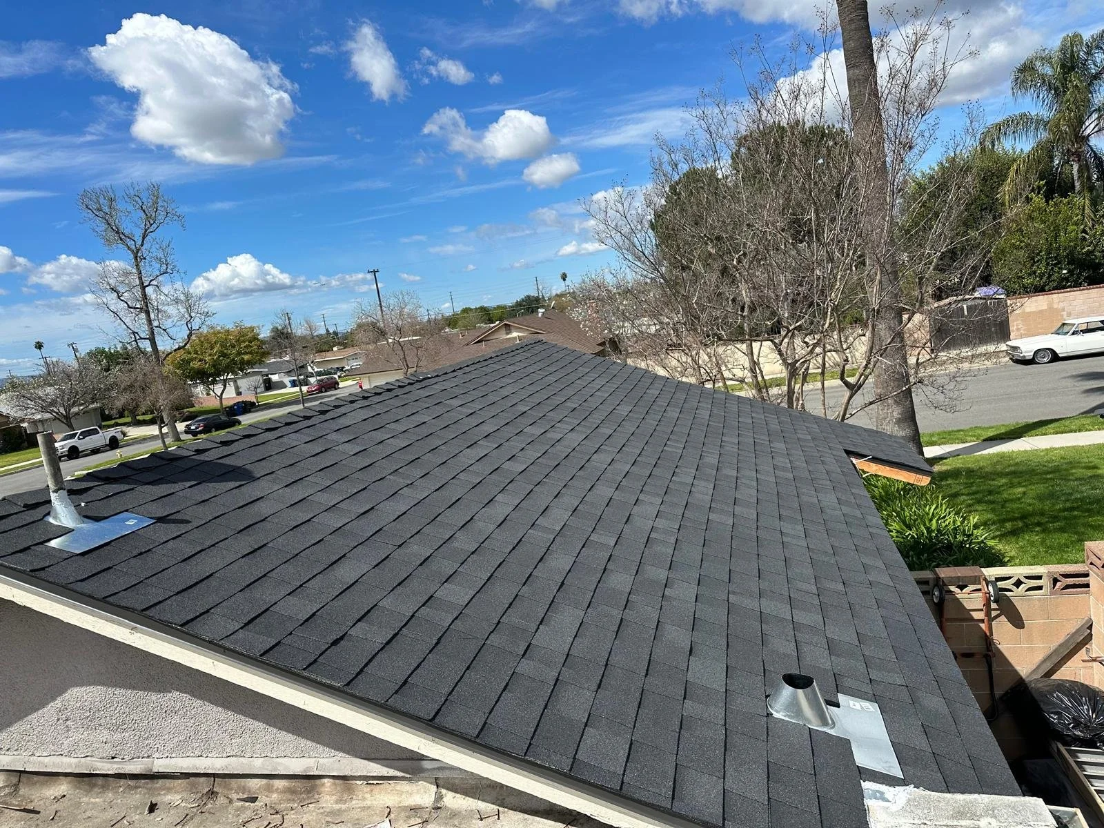 View of a residential neighborhood with a newly installed black shingle roof, partly cloudy blue sky, leafless trees, and parked cars along the street.