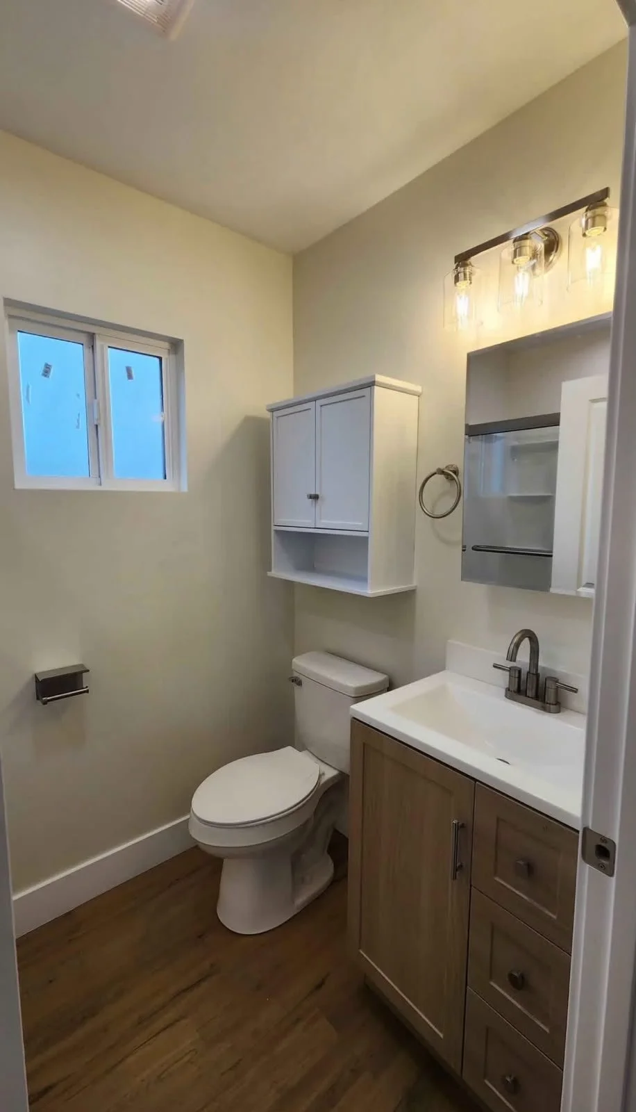 Small bathroom with a window, a white toilet, a wooden vanity with a white sink, a wall-mounted white cabinet, and a mirror with light fixtures above.