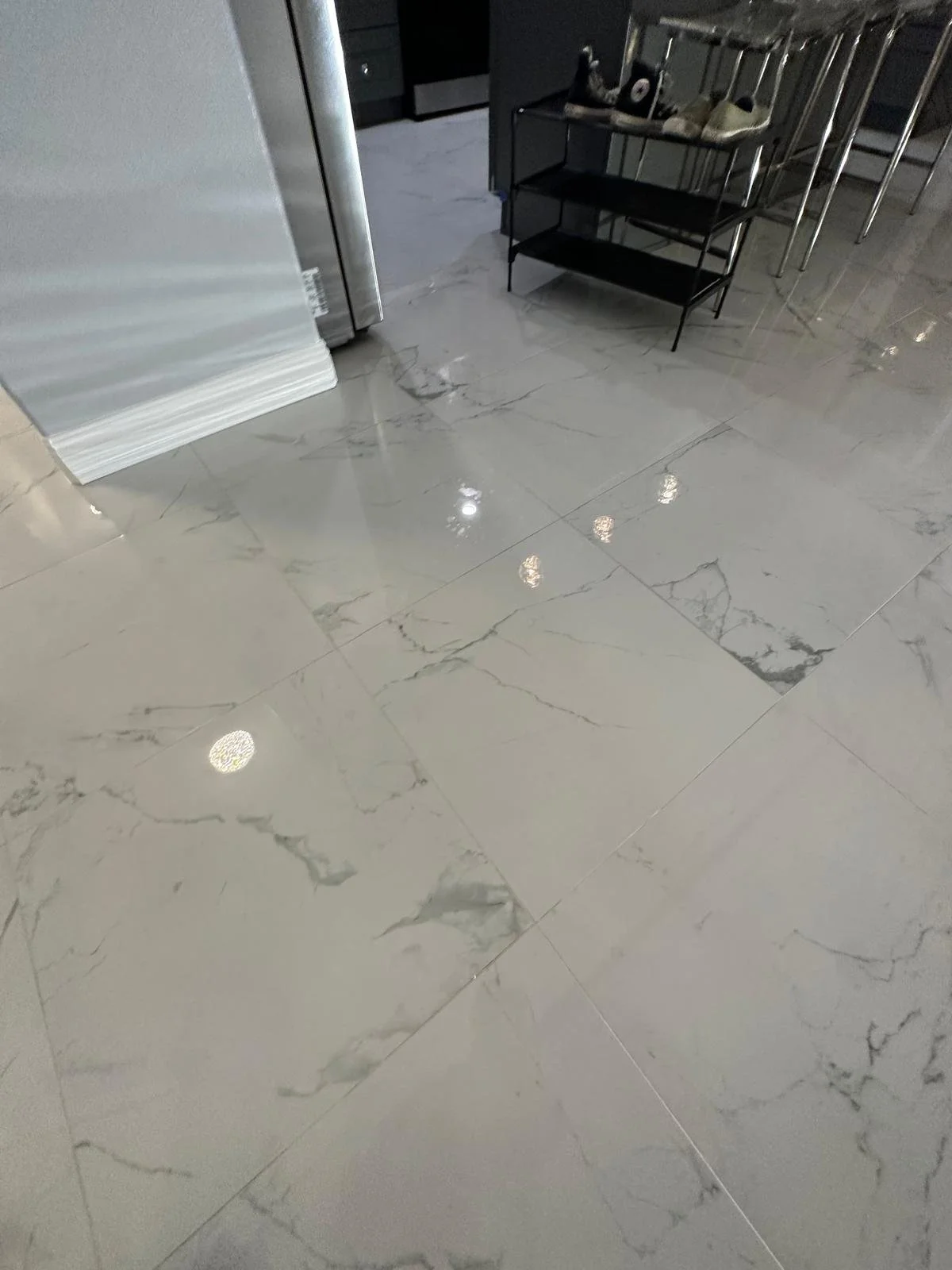 A polished marble tile floor with gray and white veining, reflecting ceiling lights. In the background, there is a metal shelf with shoes and a black and white object, beside a kitchen area with black cabinets and appliances.