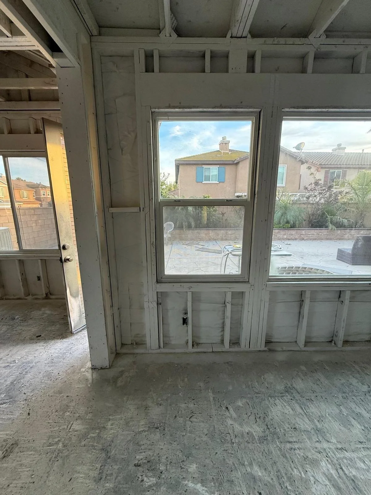 Interior view of a house under construction with unfinished walls and large windows overlooking a backyard with patio furniture and neighboring houses.
