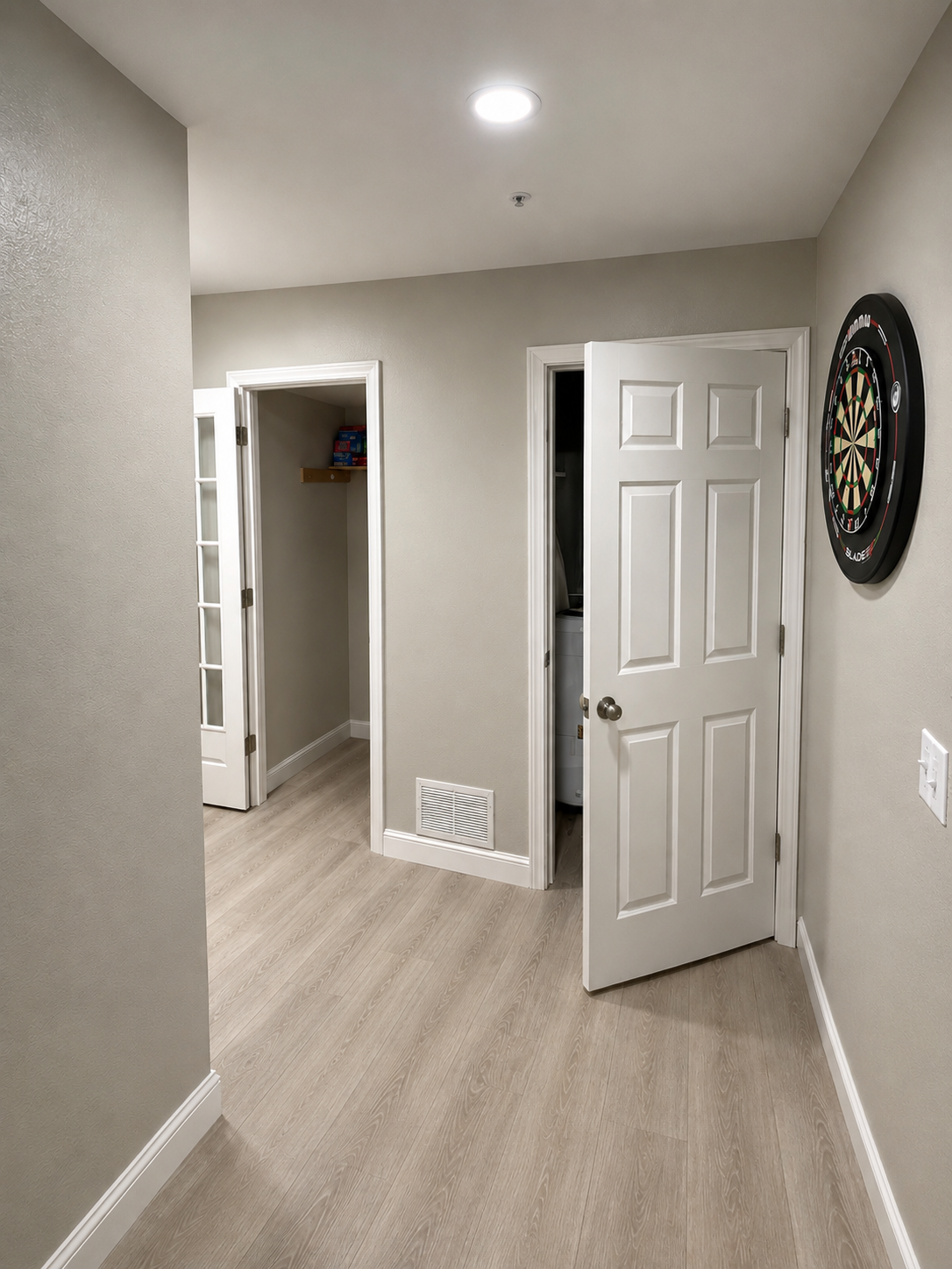 A hallway with open doors, a dartboard on the wall, and light-colored wooden flooring.