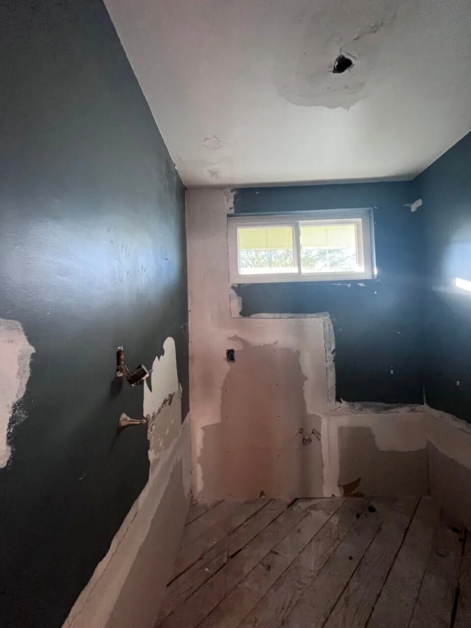 A small room under renovation with painted dark teal walls, an unfinished patch of drywall, a window at the far end, and exposed plumbing fittings on the wall.