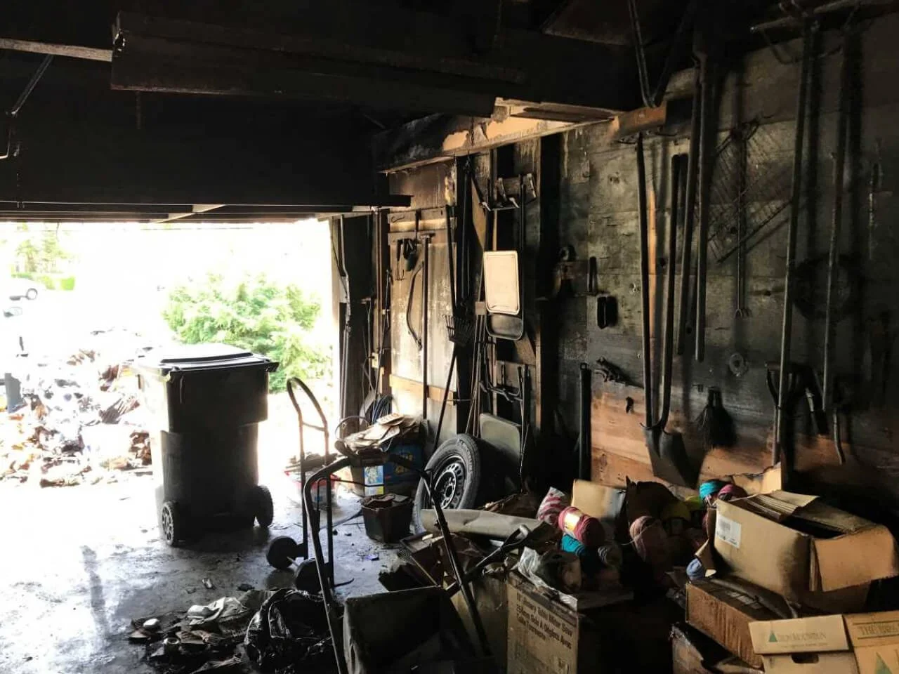 Inside of a cluttered garage with tools hanging on a wooden wall, a trash can, a box of yarn, and various scattered items on the floor.