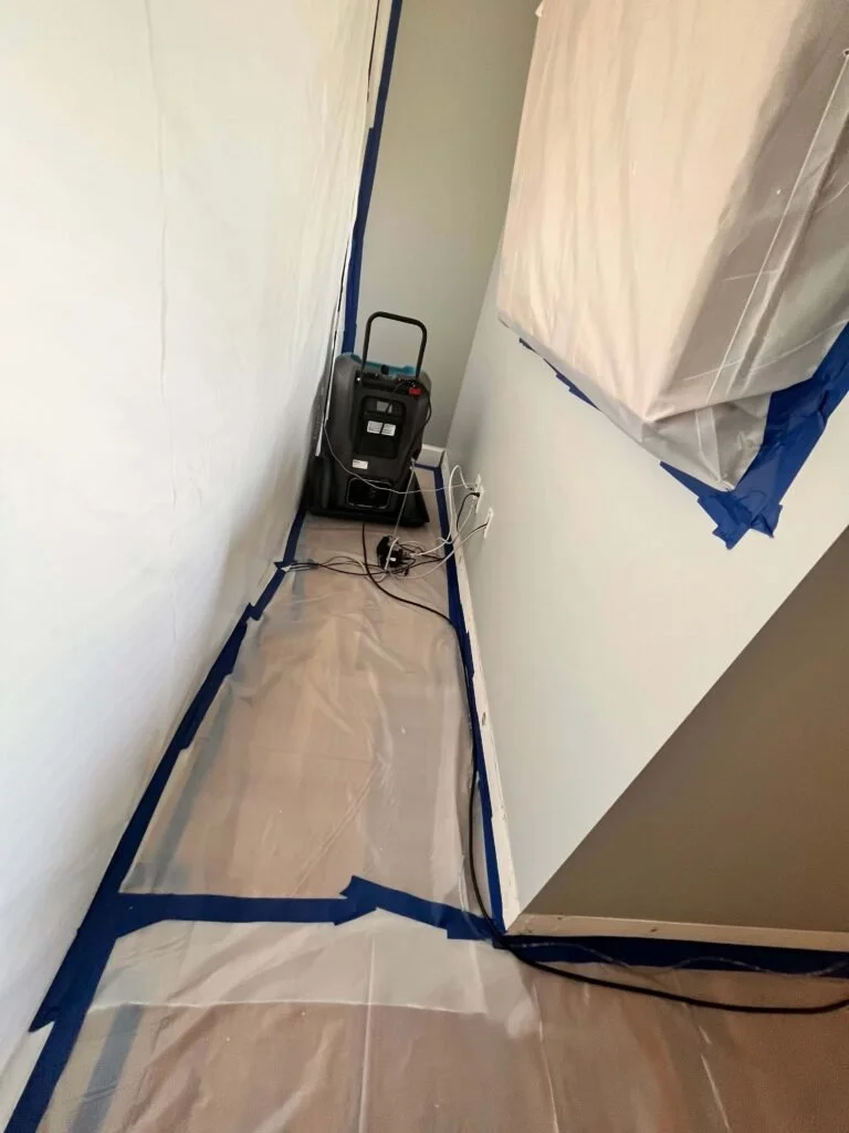A small room with walls covered in protective paper, sealed with painter's tape, and a plastic sheet on the floor. There is a portable heater or air purifier plugged into an outlet with extension cords.