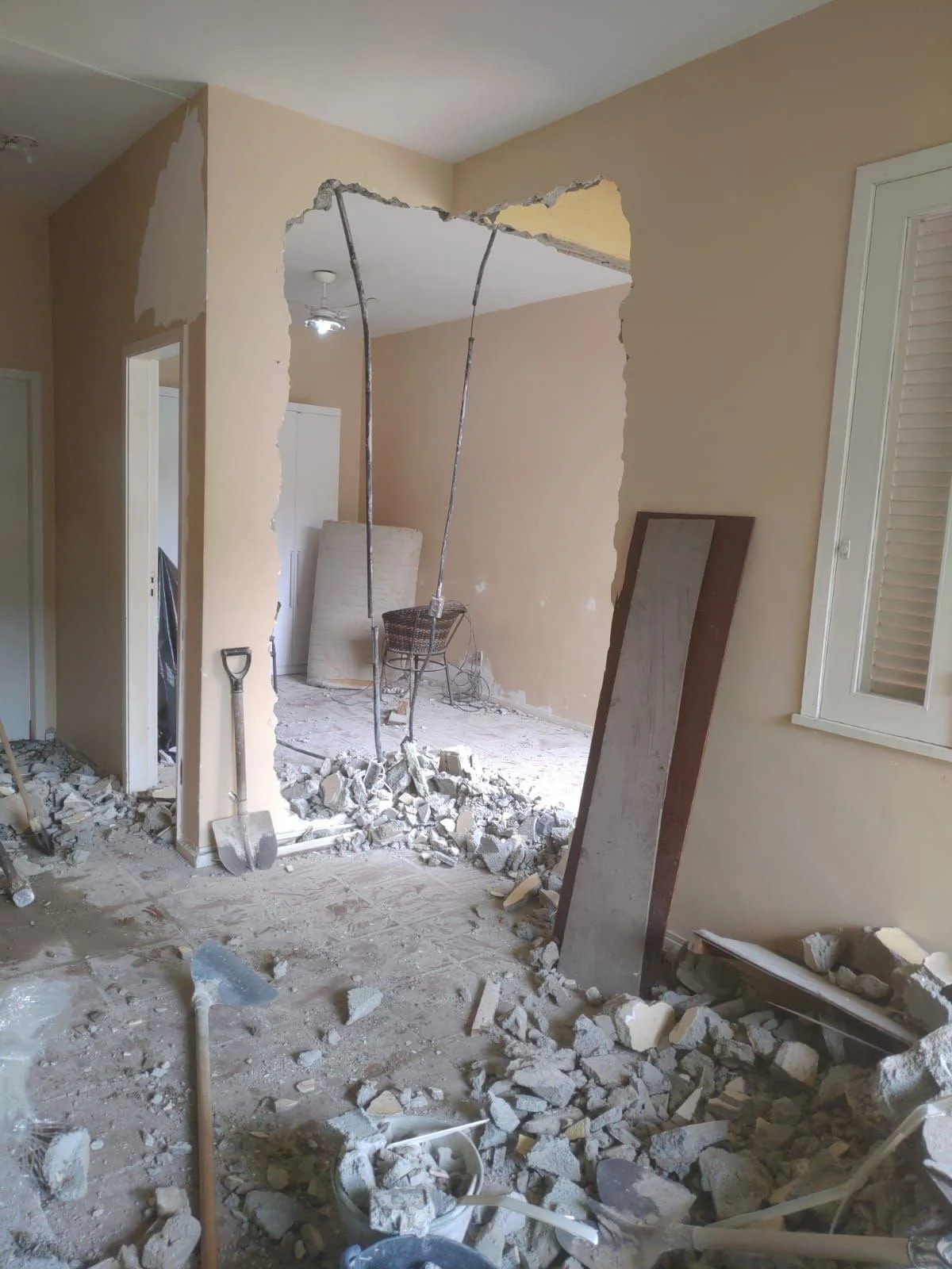 Room under demolition with debris on the floor, a partially broken wall, construction tools leaning against the wall, and a mirror propped up on a chair.