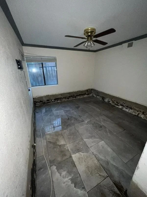 Unfinished room with peeling baseboards, a window, gray marble floor tiles, a ceiling fan, and visible damage along the walls' base.