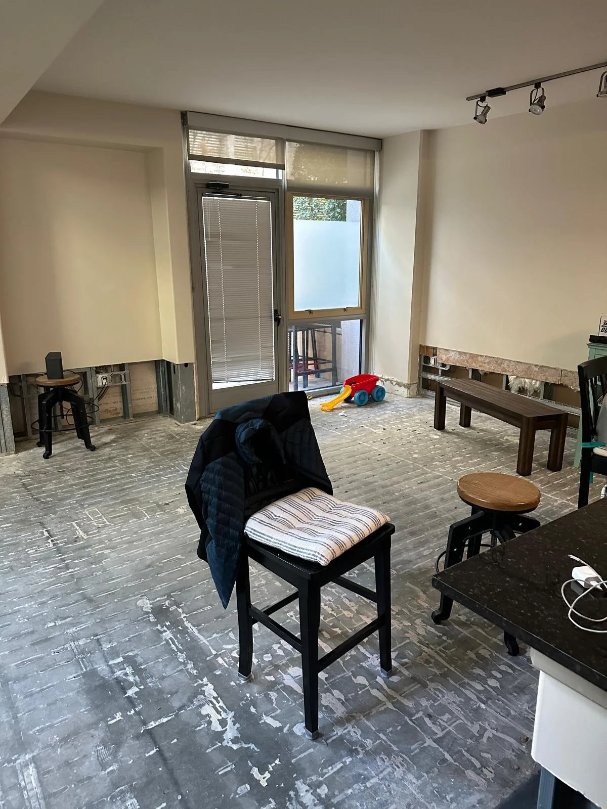 Living room under renovation with exposed flooring, a small child’s red and yellow toy wagon on the floor near the balcony door, and various furniture pieces including a black chair with a striped cushion, a small round table, and a wooden bench. The