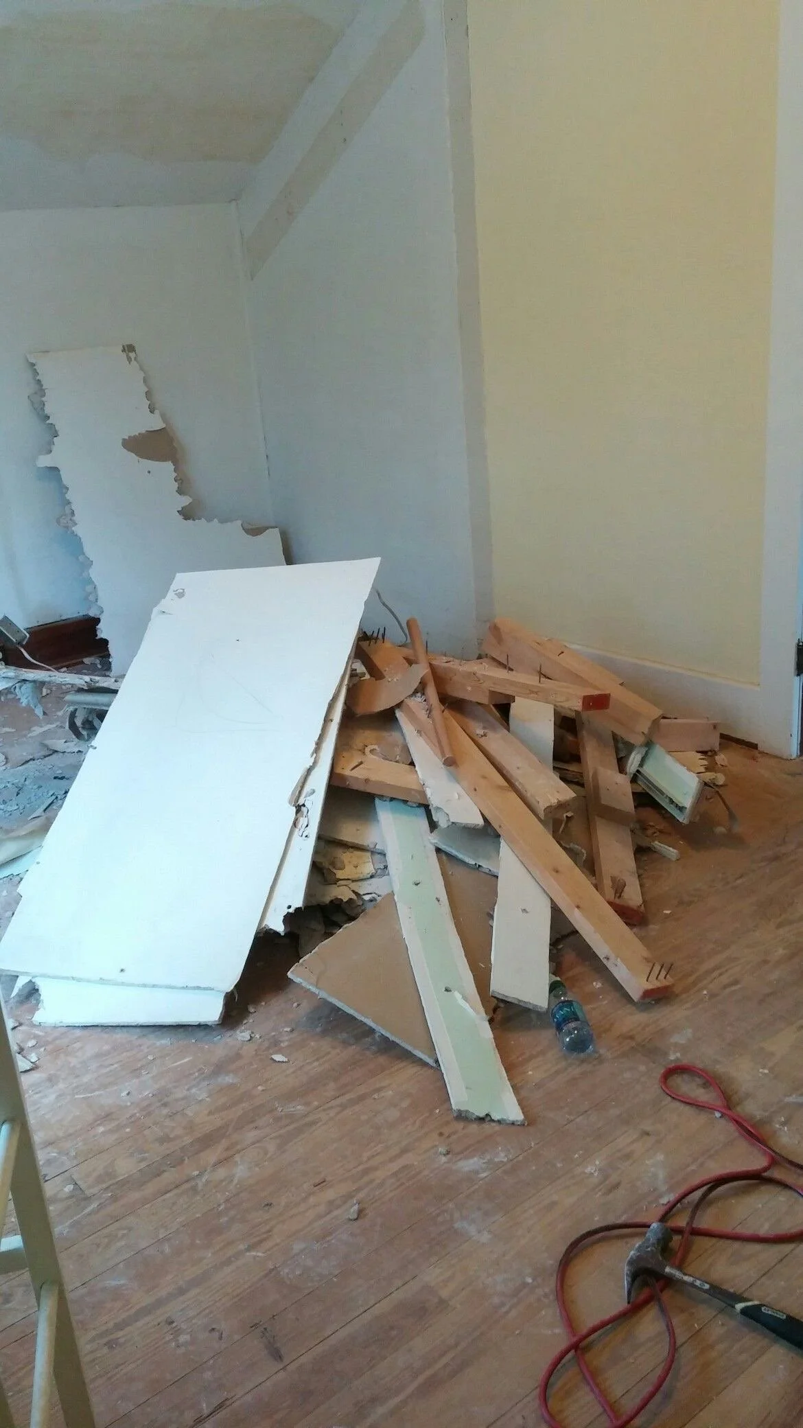 Room with debris, broken drywall, wood scraps, and a power drill on a wooden floor.