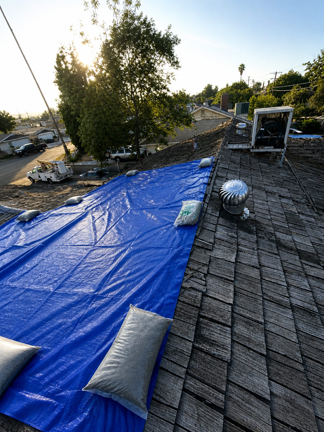 Roof being repaired or replaced with a large blue tarp secured across it, with pillows placed on top of the tarp, in a residential neighborhood during sunset.