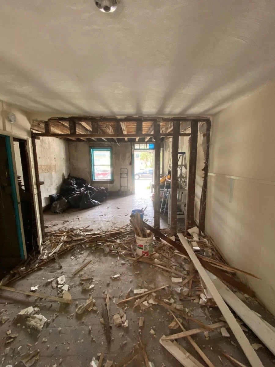 Interior of a building under renovation with scattered debris, exposed wooden beams, and construction tools. An open doorway leads outside to a street with parked cars.