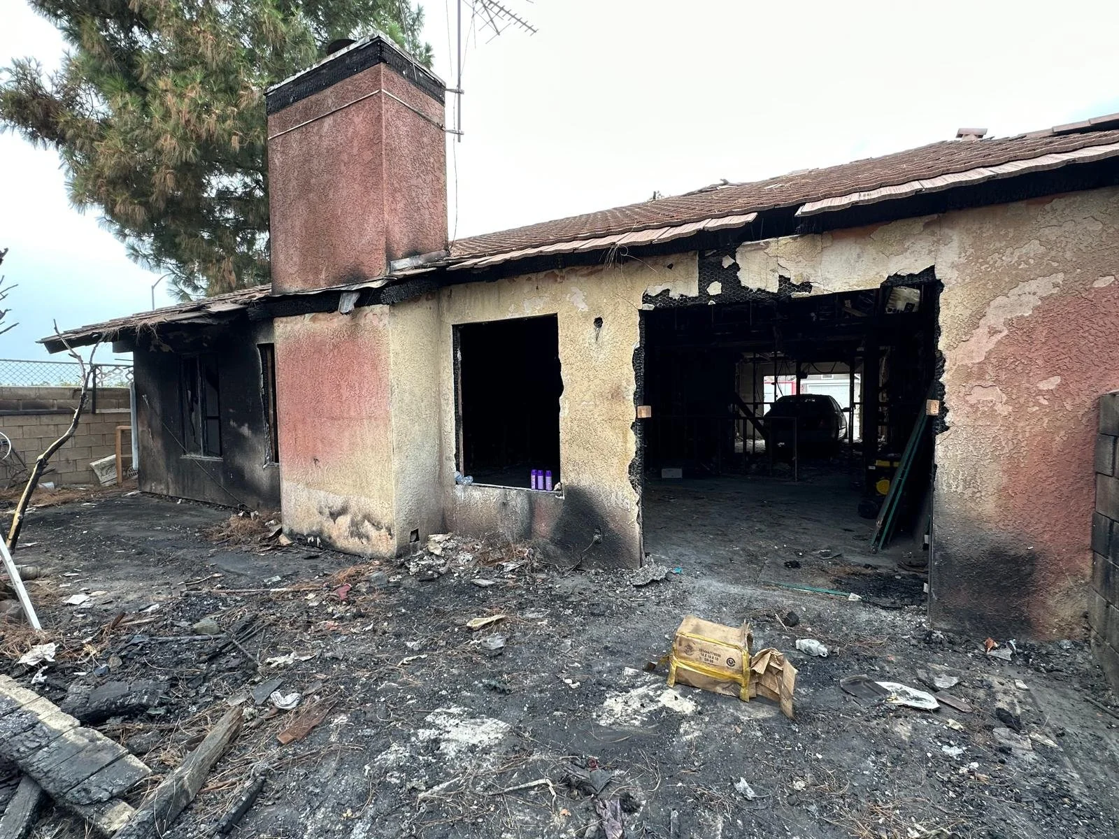 A house that was severely damaged by fire, showing blackened walls, a damaged roof, and debris on the ground.
