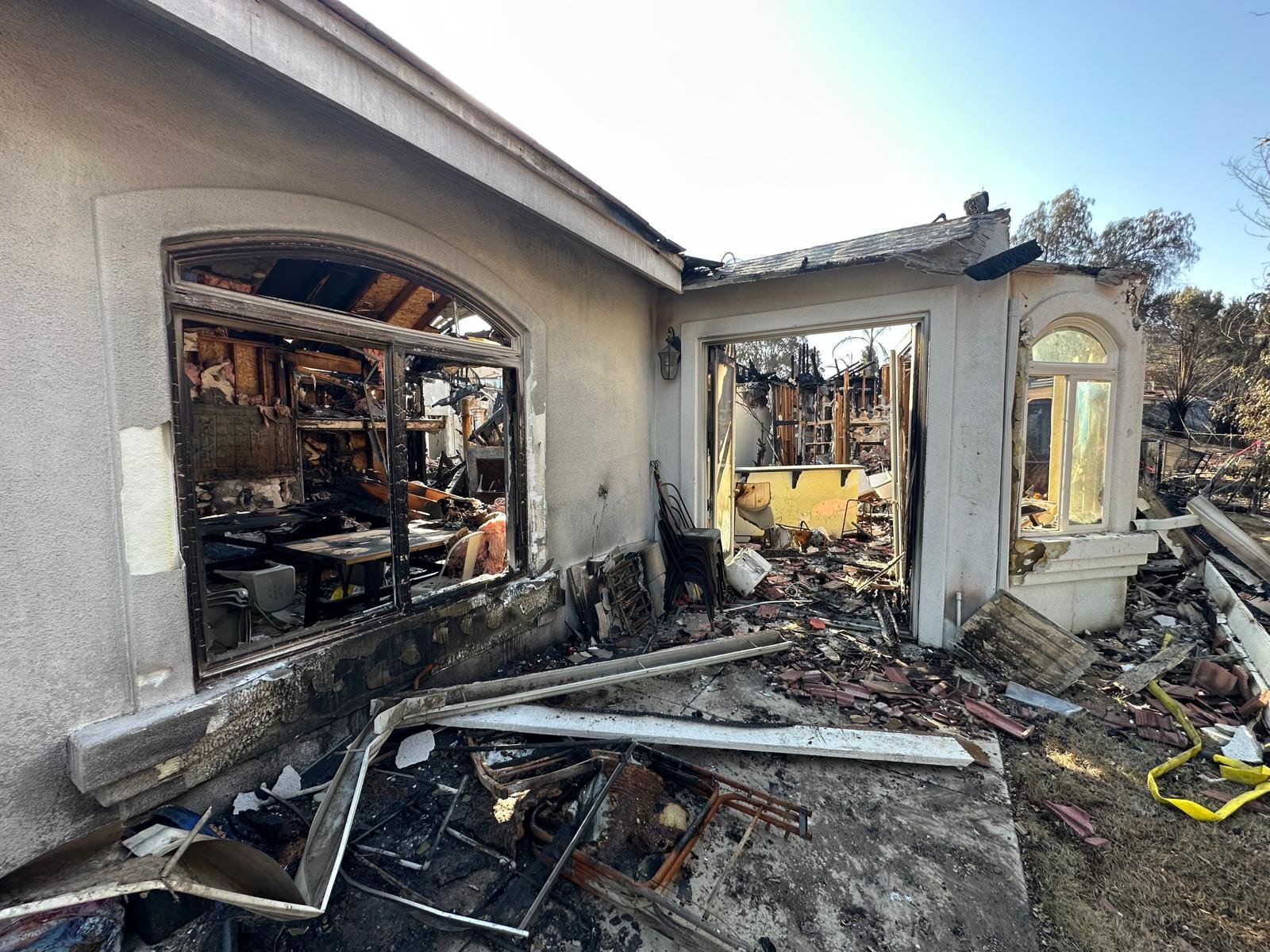 A house with severe fire damage, showing charred walls, broken windows, and debris on the ground outside.