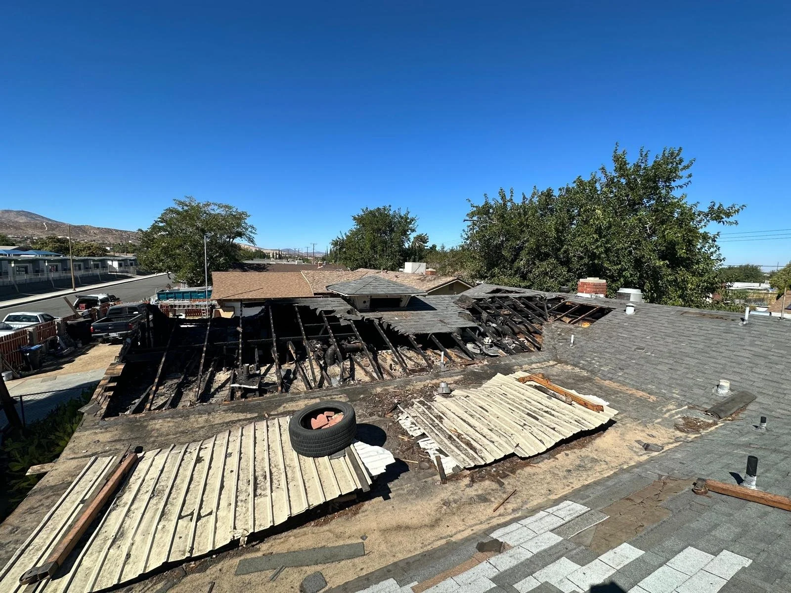 The roof of a house damaged by fire, with charred and missing sections of roofing material, exposing the roof's structure and debris scattered on the roof.