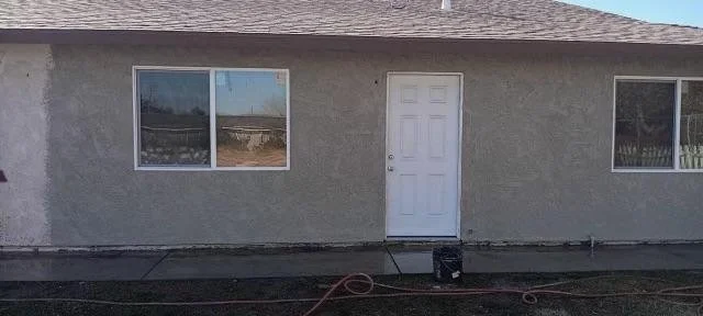 Exterior of a house with a white door, two large windows, a gray stucco wall, and a concrete patio with a hose and a small black object.