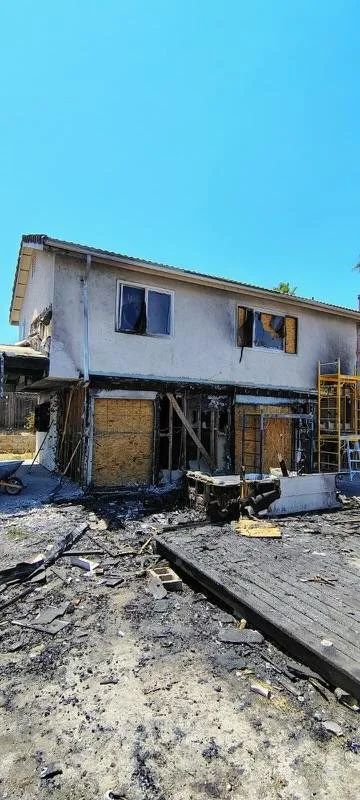 Burned and charred house with a damaged exterior, broken windows, and debris on the ground due to fire damage.