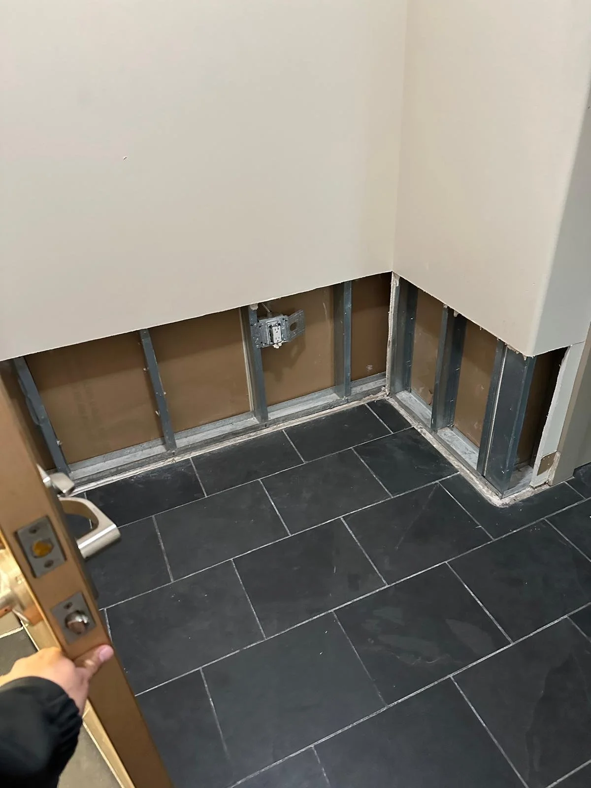 Partially renovated wall with exposed metal framing and electrical box, with black tiled floor, in a building under construction or repair.