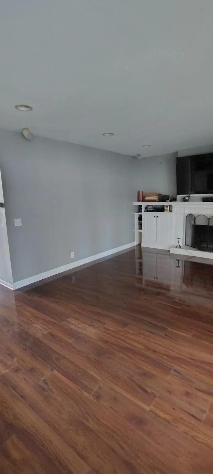 Empty living room with wooden floor, light gray walls, and a white fireplace with built-in shelves. Some books and items are placed on top of the fireplace and shelves.