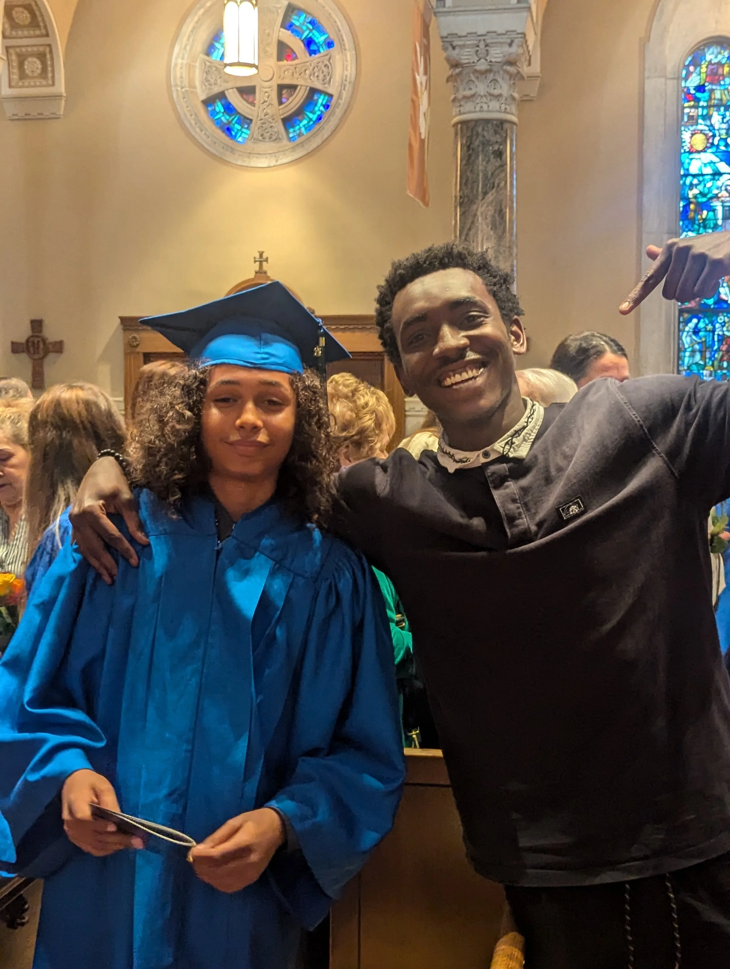 Two young adults at a graduation ceremony inside a church. The woman on the left is wearing a blue graduation gown and cap, and the man on the right is wearing a dark shirt. They are smiling and taking a selfie together, with a crowd and stained glass windows visible in the background.