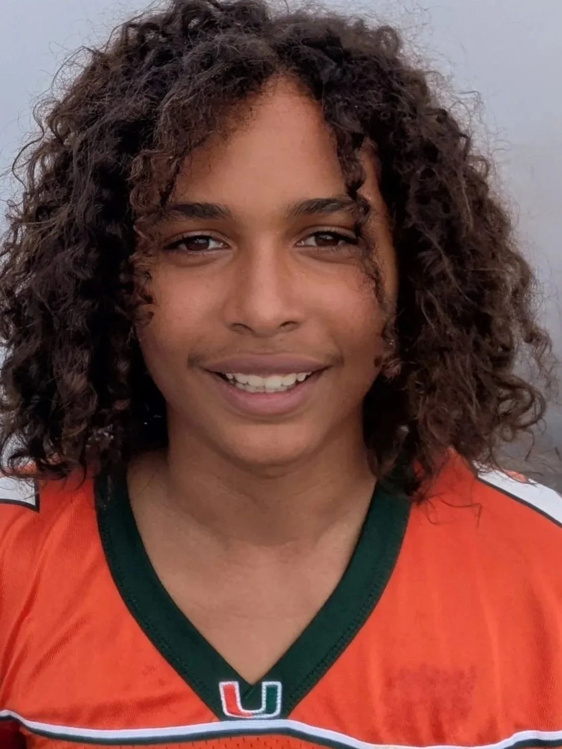 Close-up of a smiling woman with curly dark hair wearing an orange sports jersey with green and white accents.